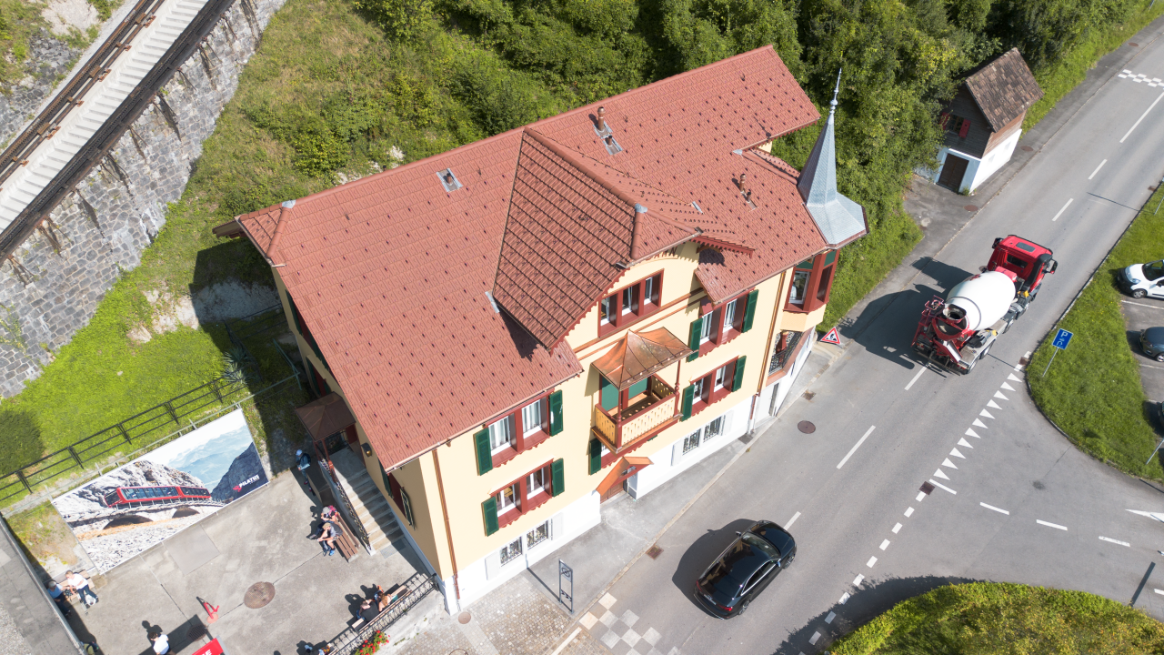 Luftaufnahme eines Hauses mit rotem Dach an einer Straßenecke, Autos und Grün im Hintergrund.
