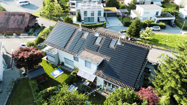 Haus mit Solarpanelen auf dem Dach, umgeben von Garten und weiteren Haeusern bei Sonnenlicht.