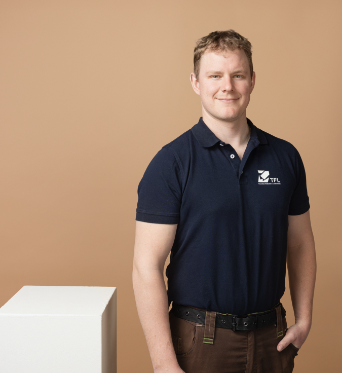Mann in dunkelblauem Poloshirt steht vor beigem Hintergrund und laechelt.