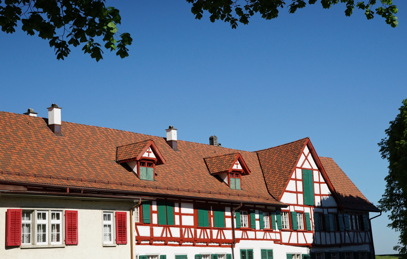 Traditionelles Fachwerkhaus mit rotem Dach vor blauem Himmel, von Baumen umrahmt.