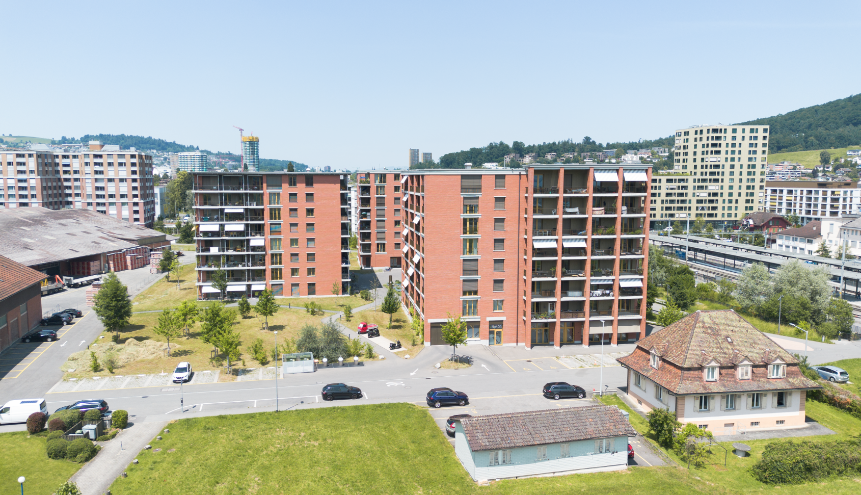 Mehrere moderne Wohnhauser mit roten Fassaden und Gruenanlagen an einem sonnigen Tag.