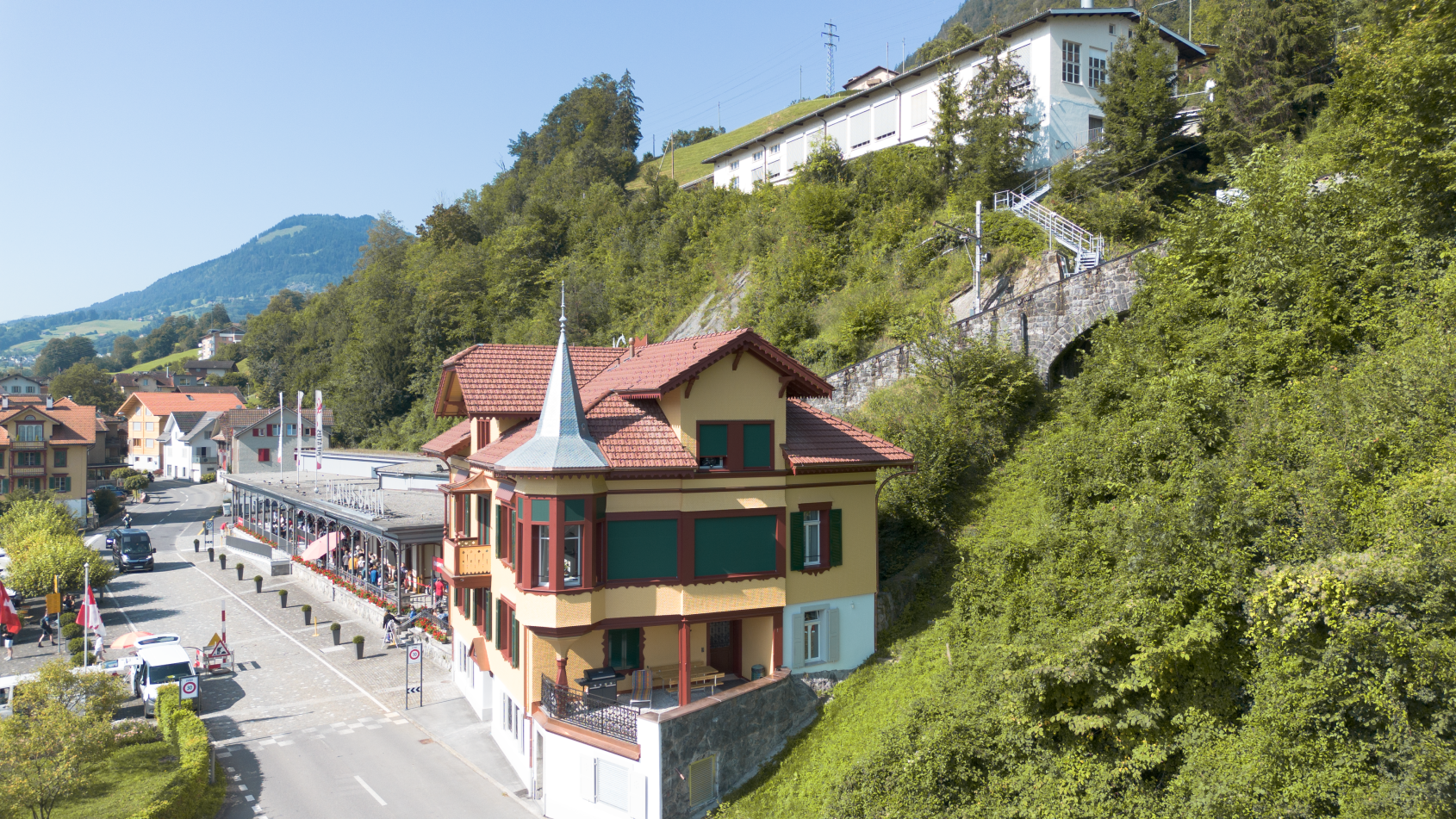 Haus mit Turm an Hang, umliegende Hauser, grune Vegetation, Berg im Hintergrund
