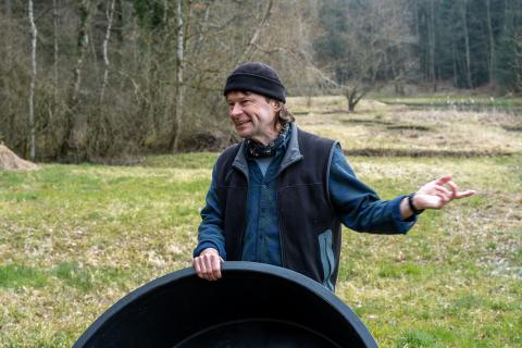 Mann mit schwarzer Muetze steht auf einer Wiese und gestikuliert mit der Hand.